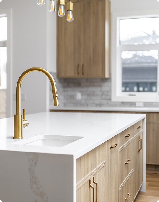 Elegant kitchen design showcasing a marble countertop with a built-in sink, complemented by a sleek brushed gold faucet. The light wood cabinetry features matching gold handles, providing a warm and cohesive look. A subtle gray subway tile backsplash adds texture to the space, while large windows allow natural light to brighten the room, enhancing the modern and luxurious ambiance.
