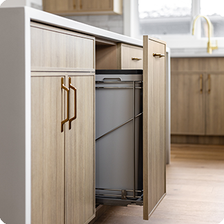 Close-up of a modern kitchen cabinet featuring a pull-out waste bin system, seamlessly integrated into light wood cabinetry with sleek gold handles. The accessory highlights the functional and stylish design, offering convenient and organized storage within a contemporary kitchen setting.