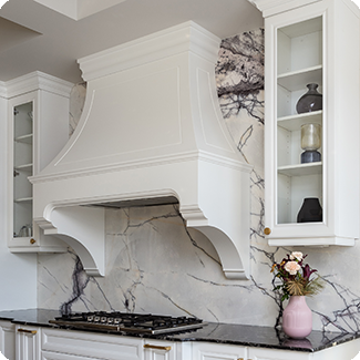 Stylish kitchen featuring a custom white range hood cover with classic molding, seamlessly integrated into a marble backsplash with striking veining. The range hood is flanked by glass-front cabinets, displaying decorative items, and a black countertop adds contrast to the elegant design. A vase with fresh flowers adds a touch of warmth and color to the sophisticated space.