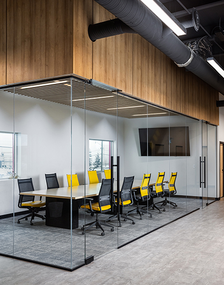 Modern office conference room with floor-to-ceiling glass walls, featuring a long wooden table surrounded by ergonomic black chairs with vibrant yellow accents. The room has a minimalist design with exposed ceiling ducts, wooden paneling, and a large wall-mounted screen, creating a professional and contemporary workspace ideal for meetings and collaborations.